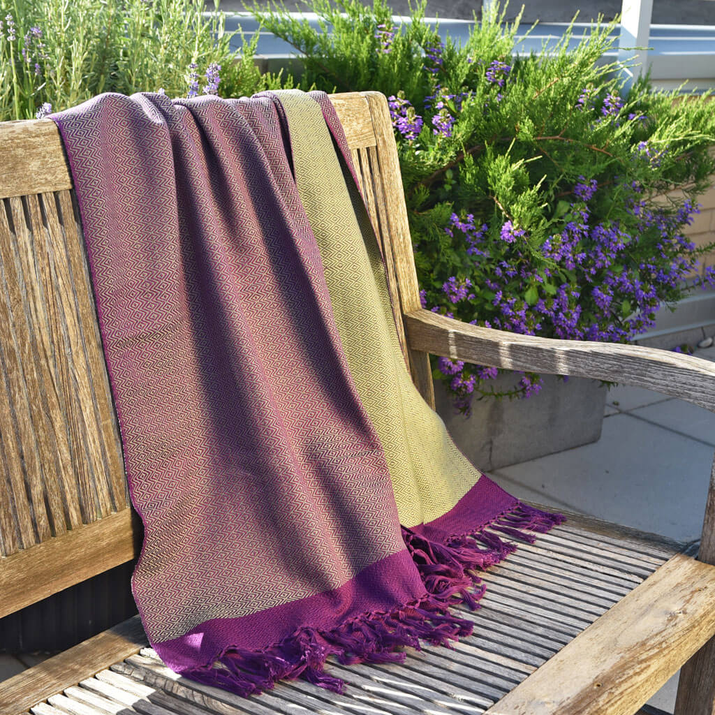 purple and sage green shawl with fringe laying over a wooden bench in front of a bush of purple flowers.